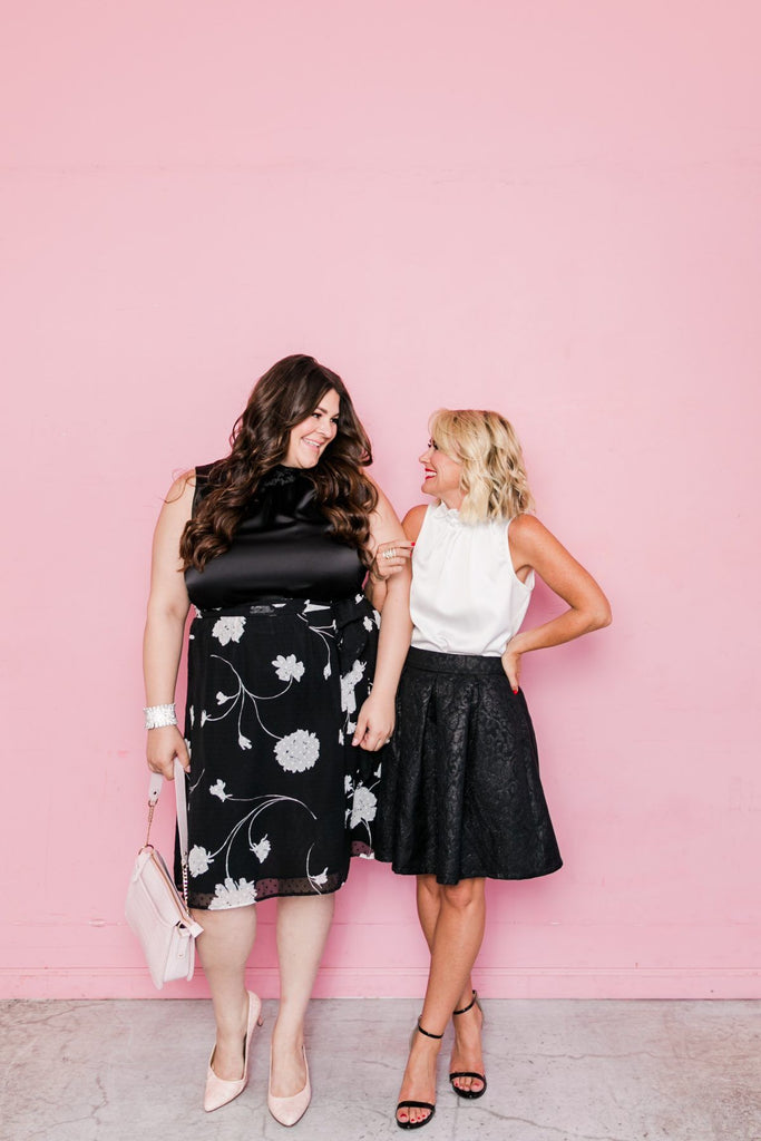 Two women wearing dresses with pink wall in the background.