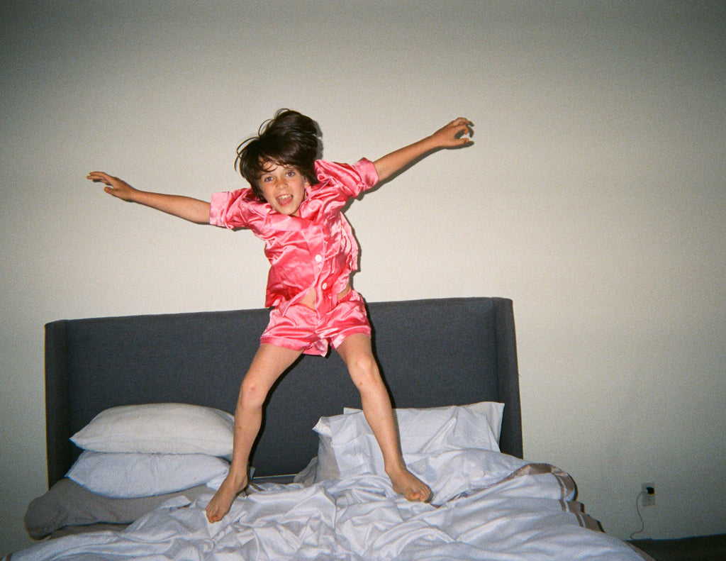 Boy in a pink button down pajama top and shorts jumping on a bed.