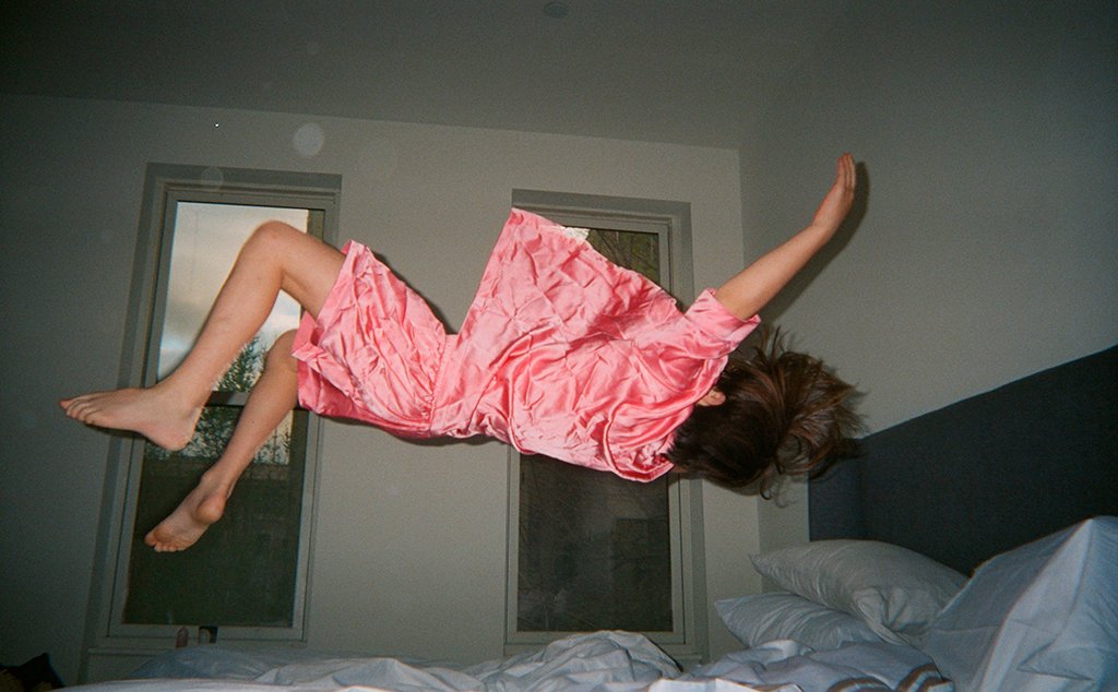 Woman in a pink silk pajama button down and shorts floating over a bed.