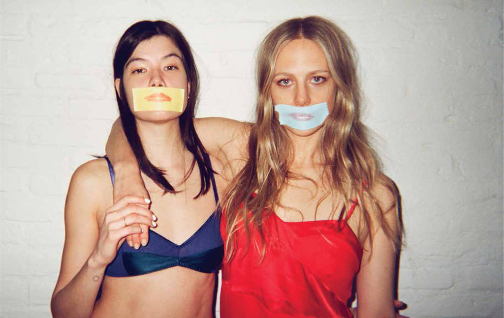 A woman in a blue bra and a woman in a red chemise with post it notes of lips over their mouths.