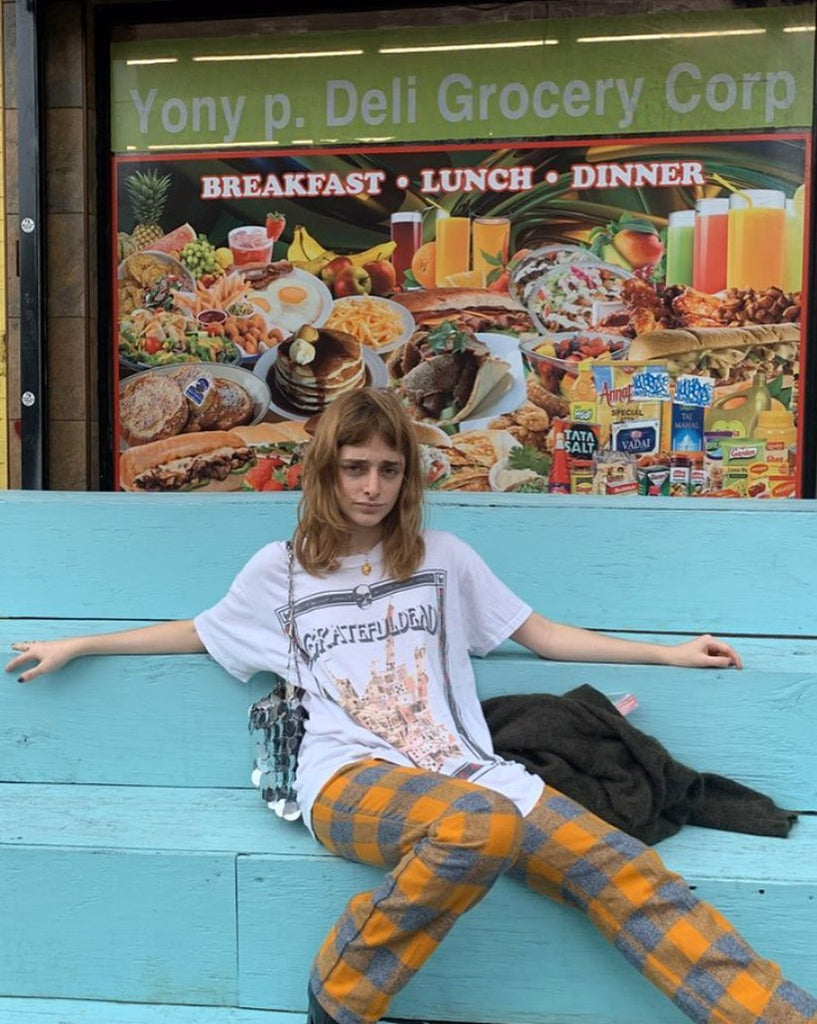 Woman in a T shirt and patterned pants sitting in front of a Deli.