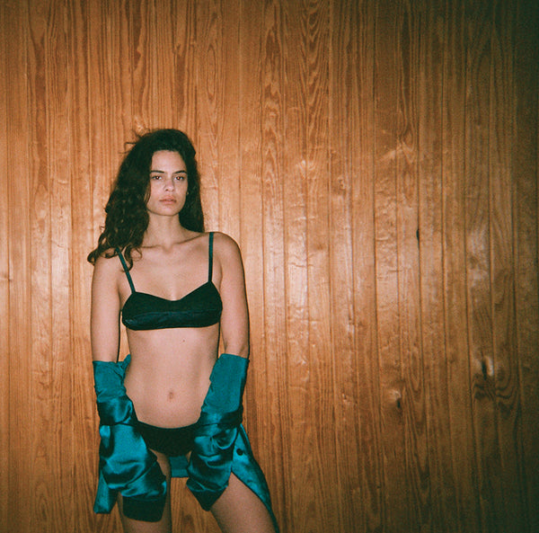 Woman in a black bra and underwear, blue gloves, standing against a wood wall.