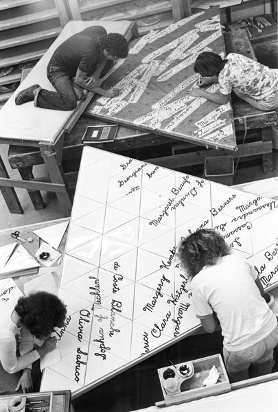Black and white photo of women painting text on a triangle sculpture.