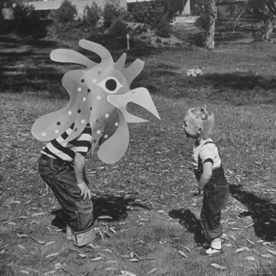 Black and white photo of a kid wearing a large paper rooster head bent over talking to another kid.