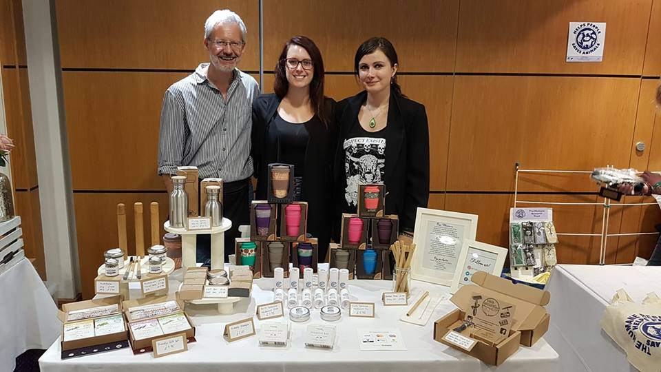 This picture shows the Full Circle Team: Paul, Emma and Johanna at their first event in Ipswich Vegan Market. They are standing behind a white table where reusable cups, plastic-free toothpaste, reusable bottles, safety razors and bamboo toothbrushes are displayed