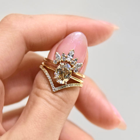 A band with three marquise shaped white diamonds with round white diamond set in yellow gold stacked above a solitaire oval champagne diamond engagement ring and a yellow gold nesting band on a light-complexion thumb. 6