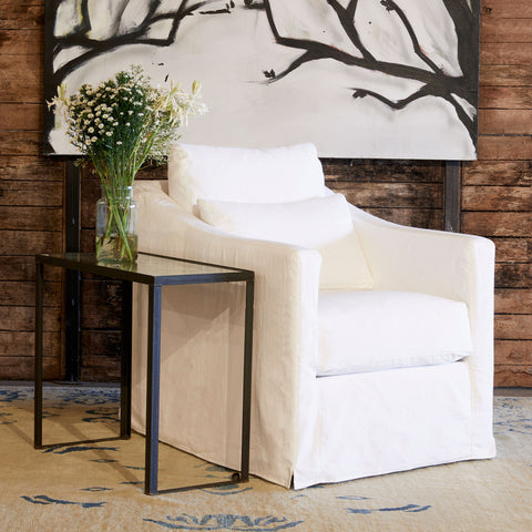 Rebecca chair in Denim White next to a glass side table. In the background is wood wall with a large painting of tree branches.