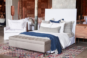 The 4月高床 is in a white slipcover, there are many decorative pillows on it, a white duvet cover and a navy blue throw. The Jaxon Chest at the end of the bed is in grey linen. To the left of the bed, there is a white slipcovered armchair. The rug is multicolored with a flower pattern. 