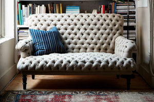  沙发是一个有很多簇绒的小物件, in a neutral color linen, 右边还有个蓝条纹枕头. 沙发在一个书架前面，前面有一块复古的蓝红地毯. 