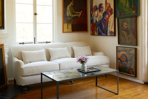 The sofa is in a neutral linen color, with 3 small lumbar pillows. It is in a small room with wood floors, in front of a window. On the right there are 5 vintage paintings, in front there is a rectangular coffee table with a vintage mirror top. There is a blue vase with purple flowers on a black tray.