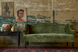  Green velvet loveseat in front of a wall with old painting next to a wood table with painted pottery 