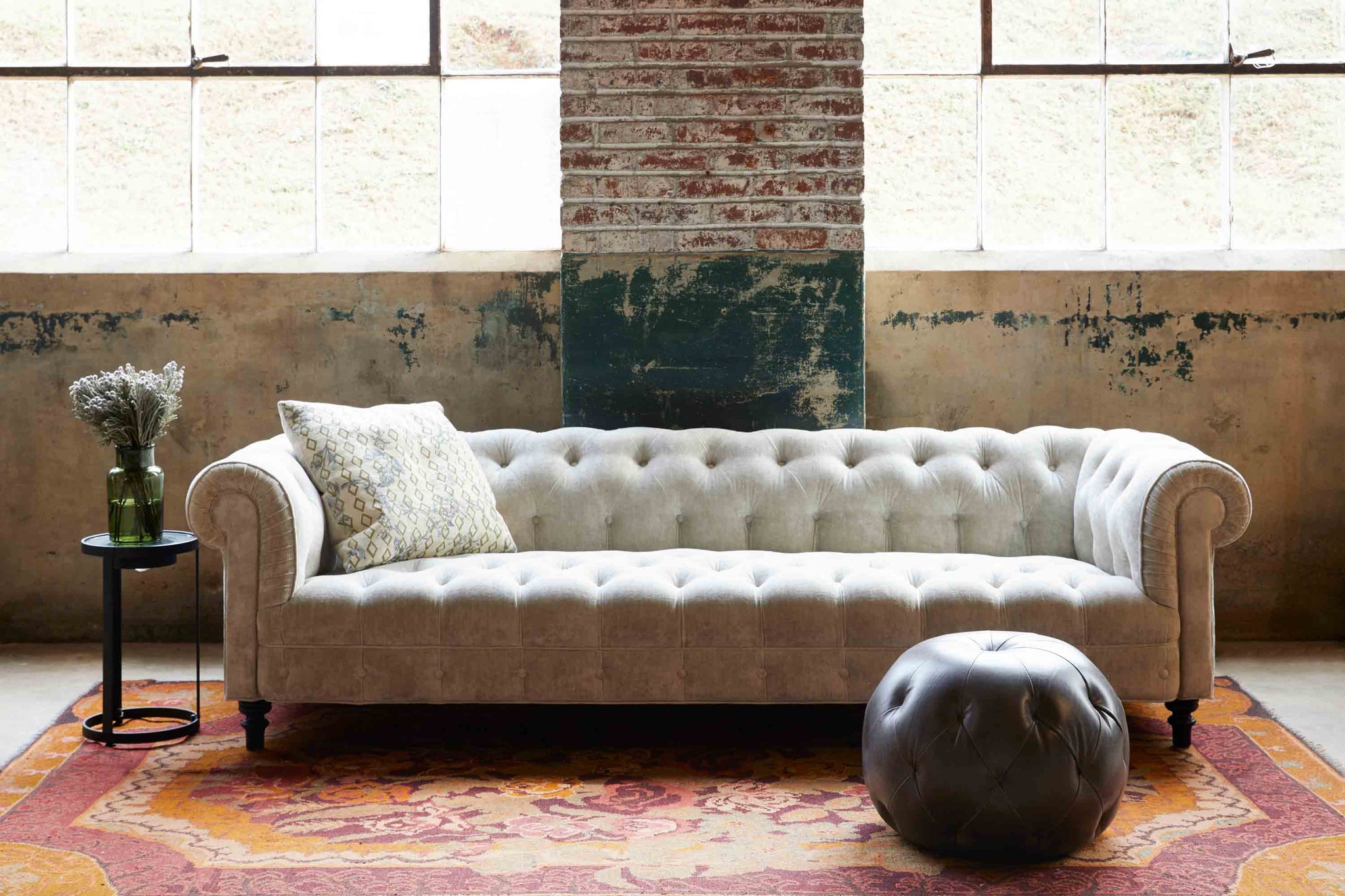  Daytime lighting of the Brook sofa in a Velluto Natural fabric, with 2 large windows and a brick wall. There is a leather ottoman in dark leather and a Rotor side table to the left. The rug has a floral pattern in the red and orange tones. 