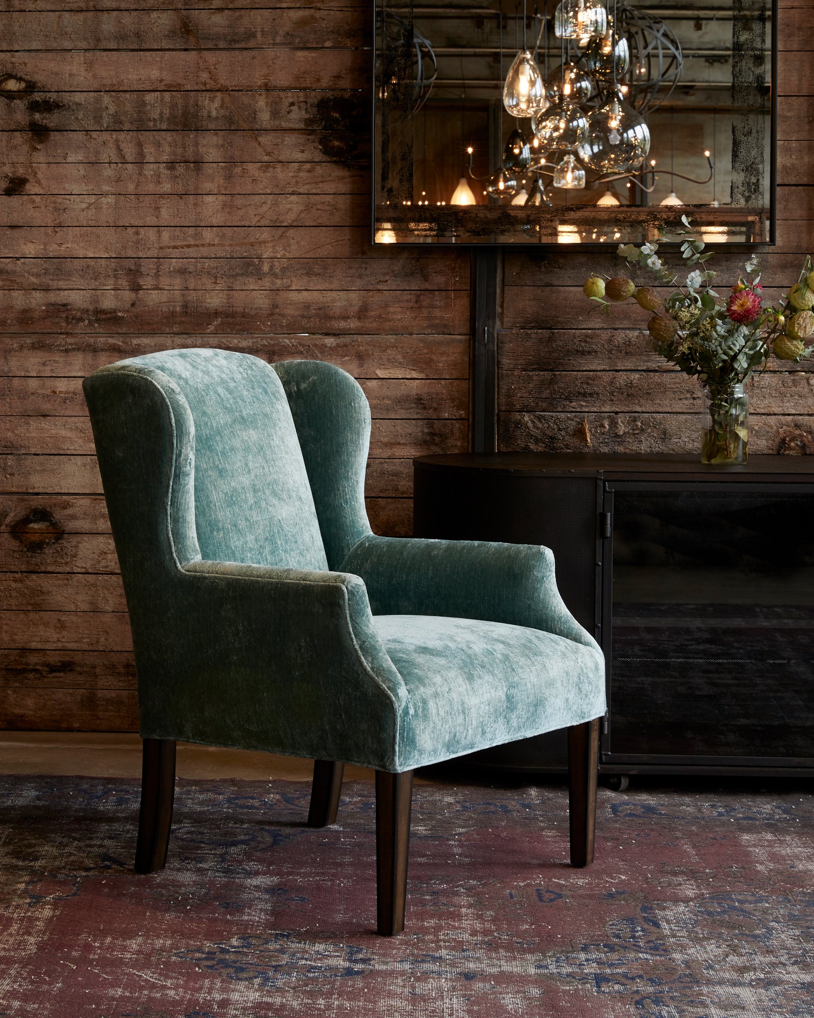 Chair in Velluto Aqua next to a dark credenza. In the background is a wood wall with a mirror and chandelier hanging. 