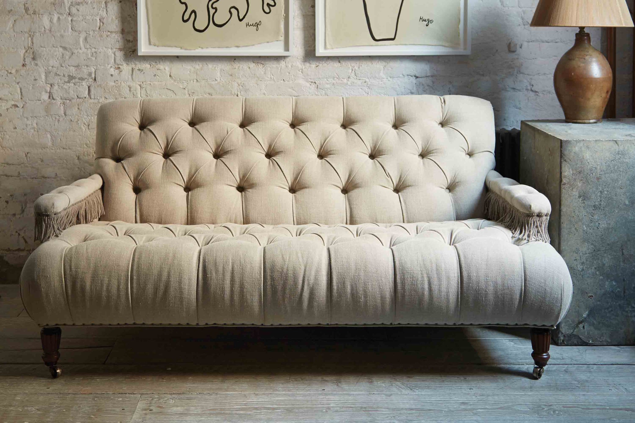  Tufted small scale sofa in a natural Vintage Flax linen with fringes on the arm rests. It is in a room wit daylight and a white brick wall and a wood floor. 