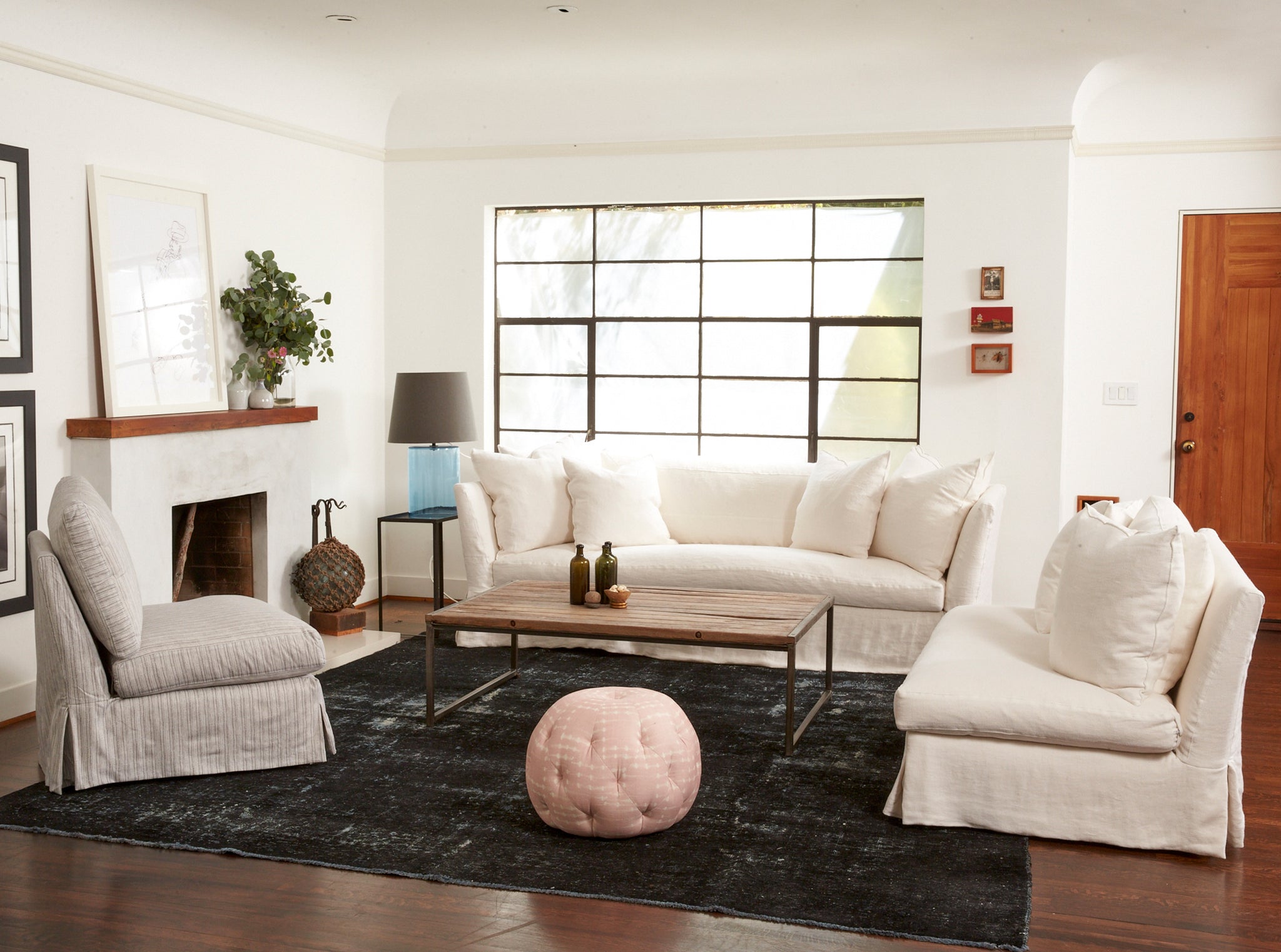  Living room setting in a white painted room with a large window. There is a dark patchwork rug with two slip covered sofas in a white fabric and a slip covered chair in a white fabric. There is a rectangular coffee table in the center of the room and a small circular pouf in a light pink fabric. Alongside one of the sofas is a side table and a cylinder table lamp on top in a light blue finish and has a dark grey fabric lamp shade.  