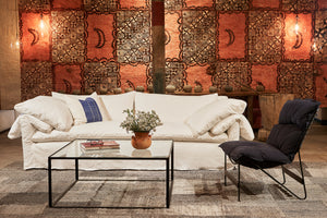  The white linen slipcovered sofa is in front of a vintage Tapa Cloth with orange and brown graphics. 右边的椅子有一个金属框架和一个深蓝色的垫子. 这张咖啡桌是金属的，上面是玻璃的. 