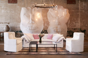  白色的沙发在一个大房间里，墙上有两个巨大的白色面具雕塑. 两边各有两把白色的沙发椅，中间有一张金属和玻璃的咖啡桌. 