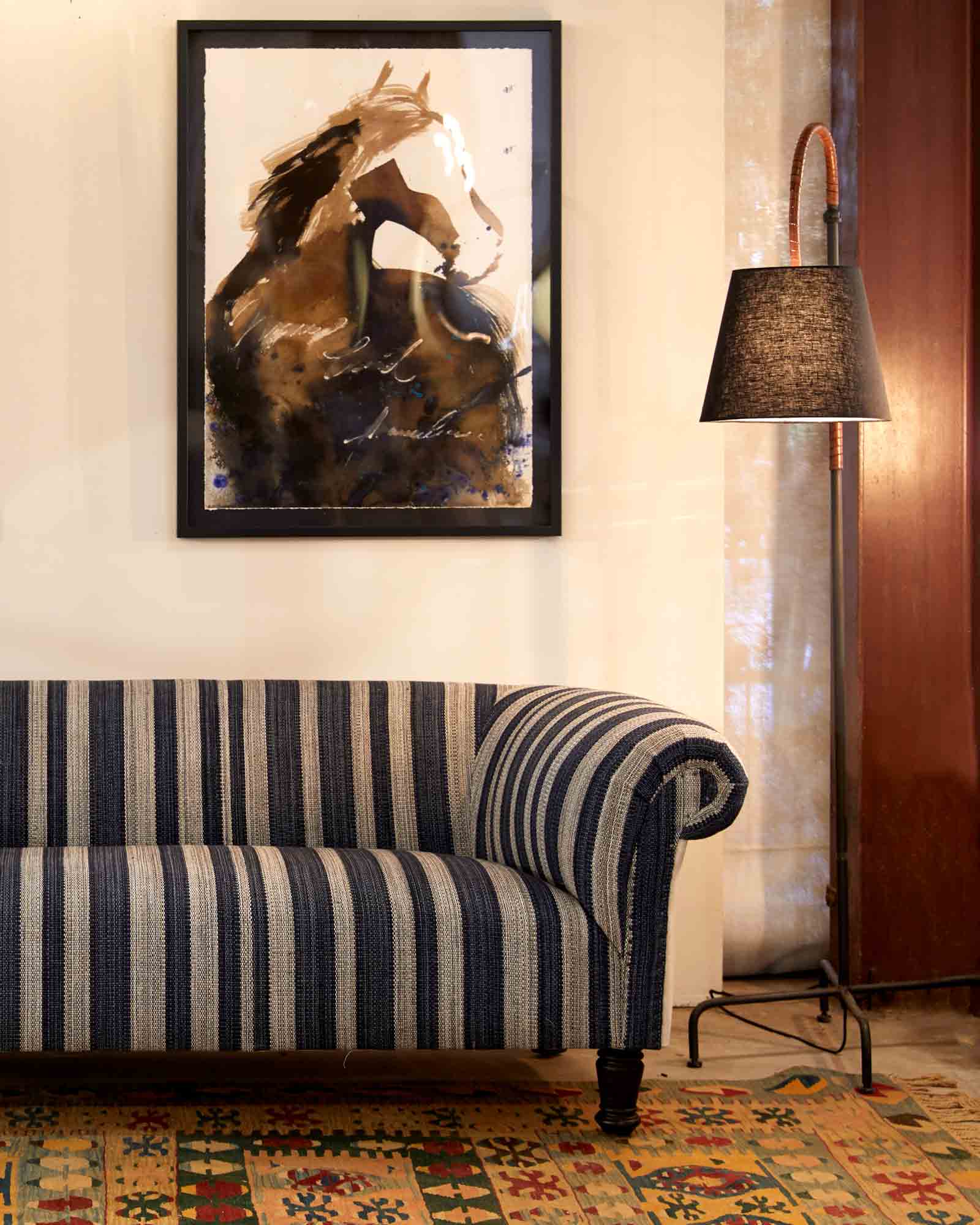  蓝色和白色织物的软垫沙发靠着白色的墙壁，墙上挂着一幅马画. 沙发旁边是金属底座落地灯，有一个深棕色的灯罩.  