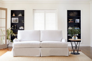  丹宁白(Denim white)的白色沙发套在自然采光的客厅里. 后面墙上有一扇窗户和两个黑色的书架. 木制和金属的圆边桌上有一个绿色花束的花瓶. 