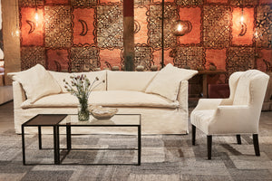  哈瓦那沙发在一个天然的奶油色织物. 坐在前面的老式塔帕布上，有展厅照明. 咖啡桌是金属和玻璃的，上面有一个碗和鲜花. 右边是约翰·德里安(John Derian)设计的浅色面料的蝴蝶椅. 