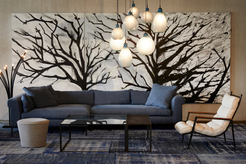 Henrietta 2pc Sectional in Rye Midnight Blue next to a glass coffee table, a light ottoman and a light colored chair. Underneath the pieces os a patterned blue rug. Behind the sectional is a large painting of tree branches. Above hangs multiple jug lamps. 