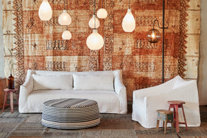  在布里瓦德象牙的Tombo椅子旁边的白色沙发, 两个木头凳子, 一盏深色落地灯和一个有图案的奥斯曼. 背景是一幅大型图案画. 在所有部件的上方悬挂着照明部件. 