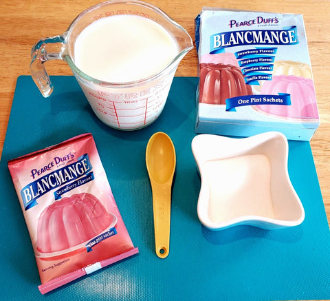Blancmange packet on a blue cutting board with 1 TBSP, sugar filled dish, and a measuring cup filled with milk