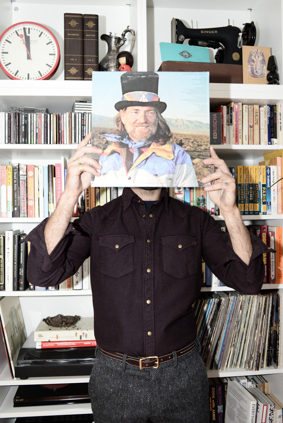 Dylan shot in front of a bookcase holding a Willie Nelson record over his face. He is wearing a burgundy western shirt.