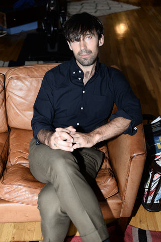 Dylan seated on a leather couch wearing a black button down shirt and green suit pants.