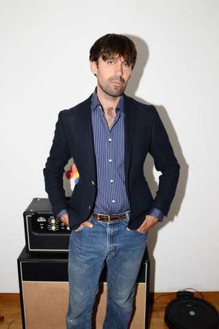 Full length shot of Dylan wearing jeans, a blue and white striped dress shirt, and a navy blazer in front of a guitar amp.