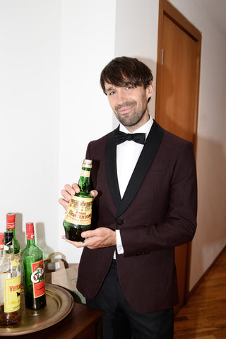 Dylan holding a bottle of amaro wearing a burgundy dinner jacket.