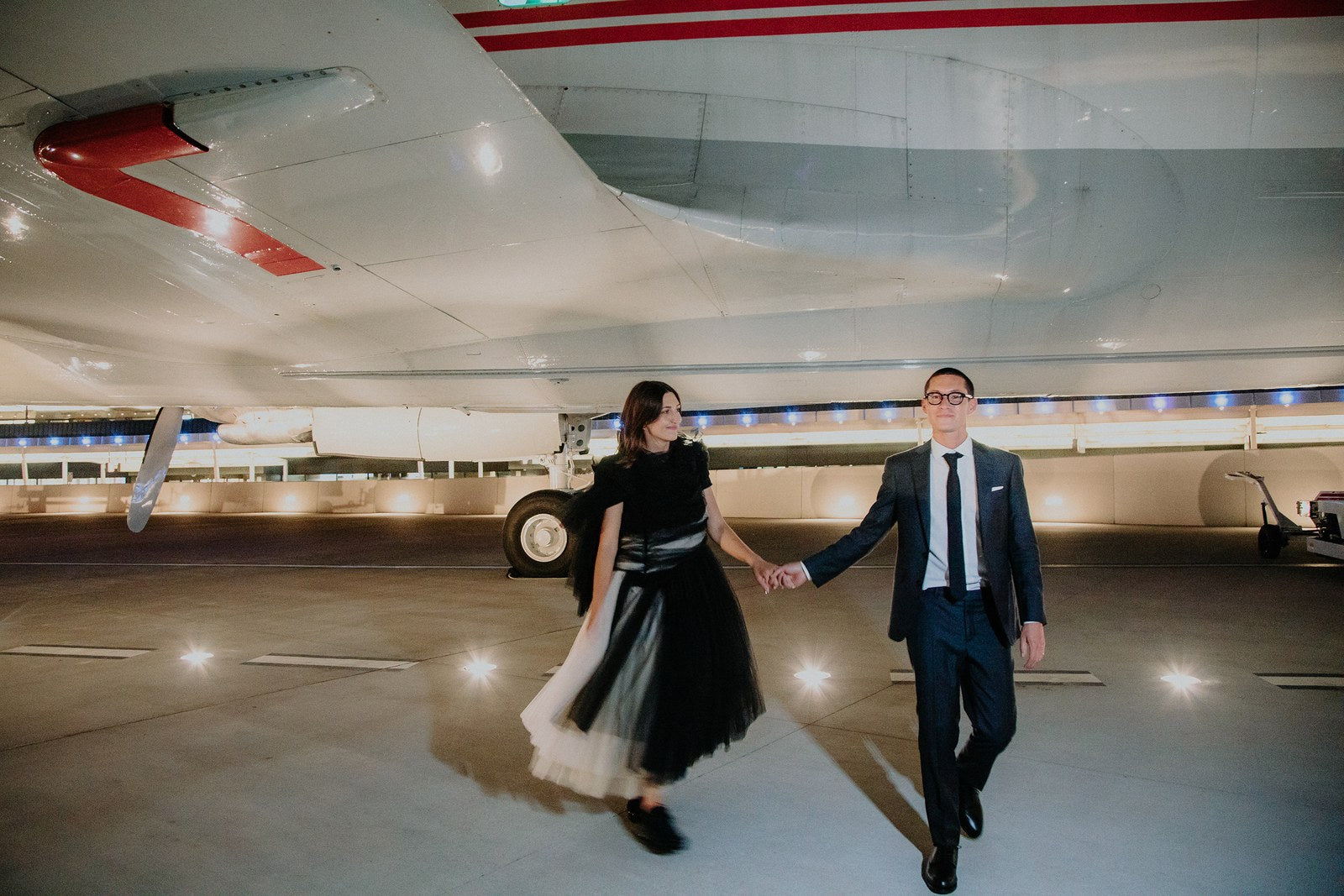 Wedding photo of Perry and Adri in front of an airplane, holding hands. Perry wears a custom Brooklyn Tailors suit.