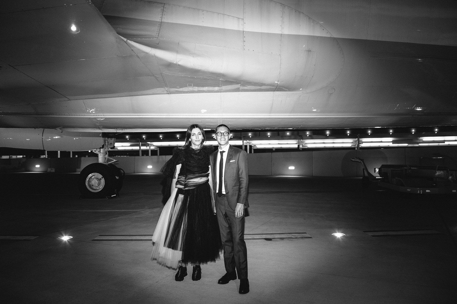 Black and white image of Perry and Adri in front of an airplane at their wedding. Perry wears a custom Brooklyn Tailors suit.