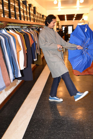 Model wearing a gray rain coat and navy pants. He is holding a bright blue open umbrella in a clothing store.
