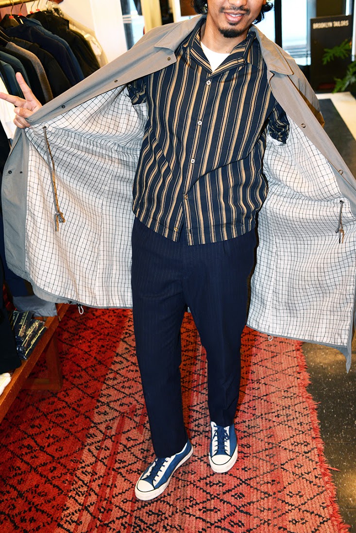 Model wears navy striped dress trousers, a navy and tan striped shirt jacket and a gray rain jacket opened to show the white and gray grid pattern lining on the inside. 