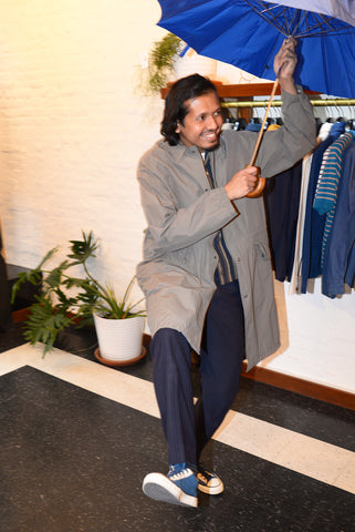 Model is wearing a gray rain coat opened over a navy and tan shirt jacket with navy pants and sneakers. He is holding an open bright blue umbrella in a clothing store.