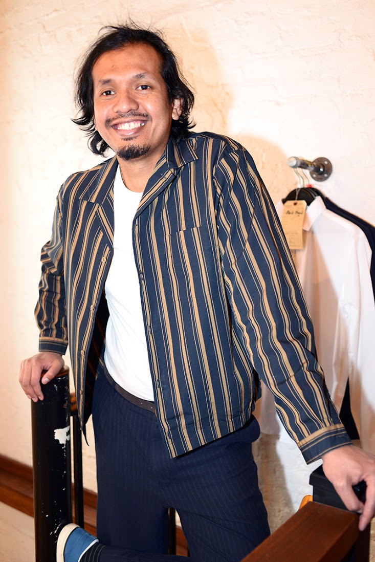 Model wears navy striped dress trousers, a brown suede belt, a white t-shirt, and a navy and tan striped shirt jacket.