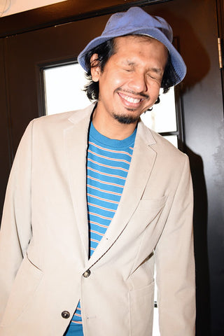 Supplemental shot of model wearing sand colored unstructured suit with blue and red striped t-shirt and bucket hat. He is squinting. 