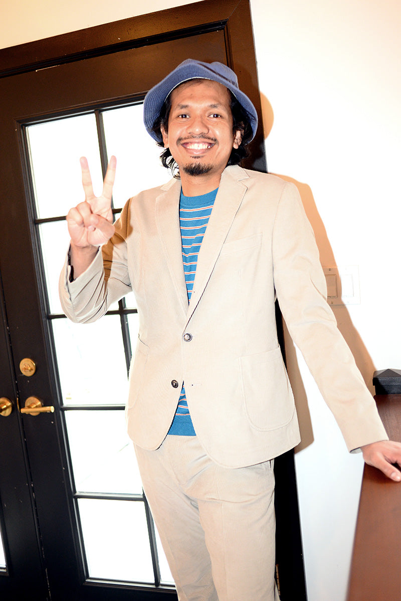Model is wearing a sand colored unstructured suit with a blue and red striped t-shirt and a blue bucket hat. He is giving a peace sign. 