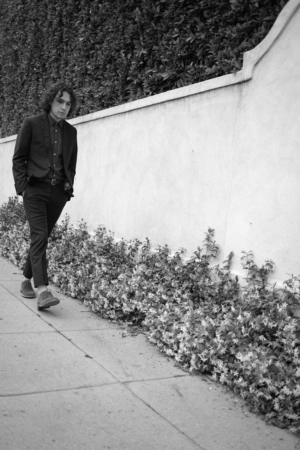 Black and white image of model walking in front of a wall, wearing a dark suit and button down shirt.