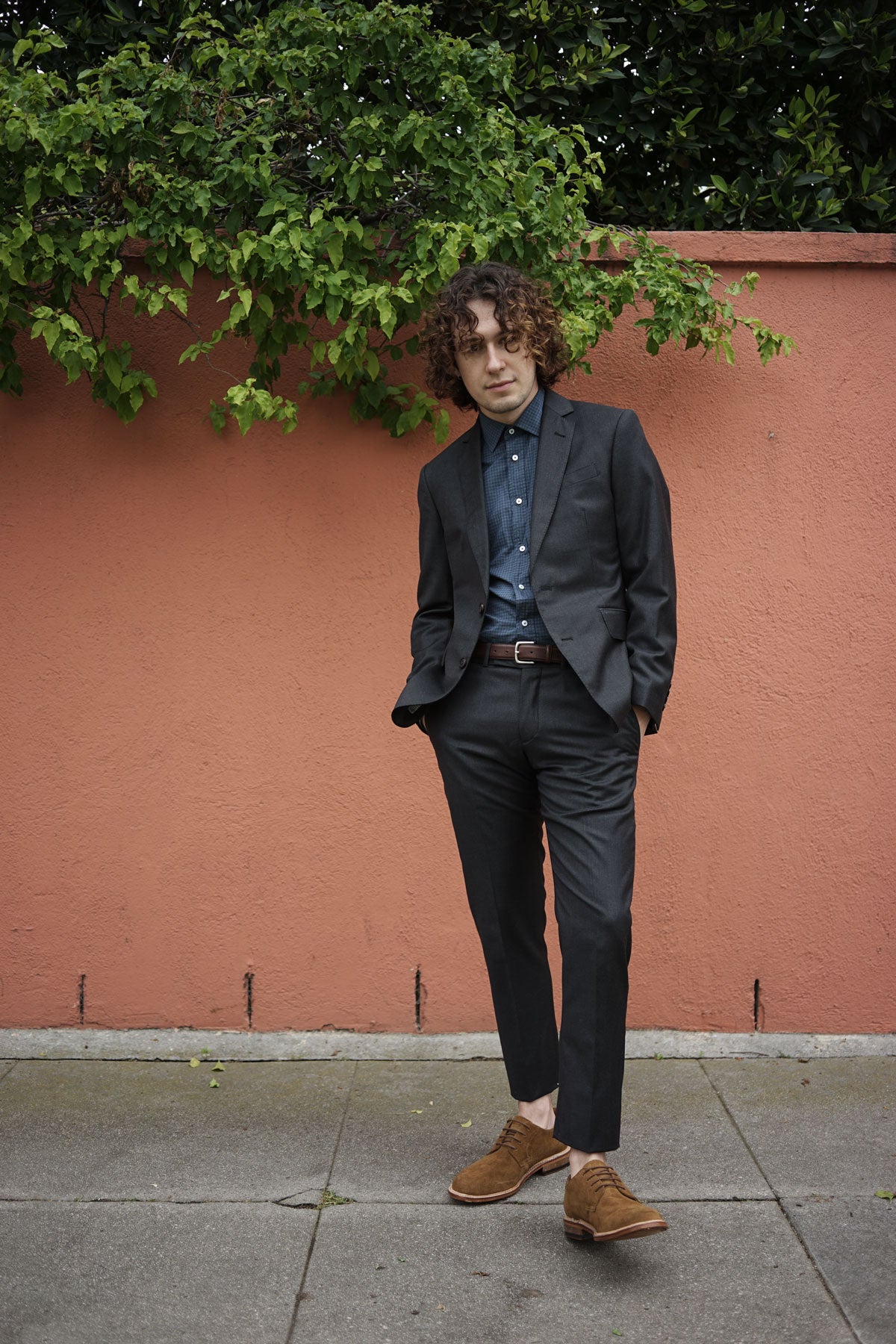 Model wears black suit with blue check shirt and brown leather belt in front of a terra cotta wall.
