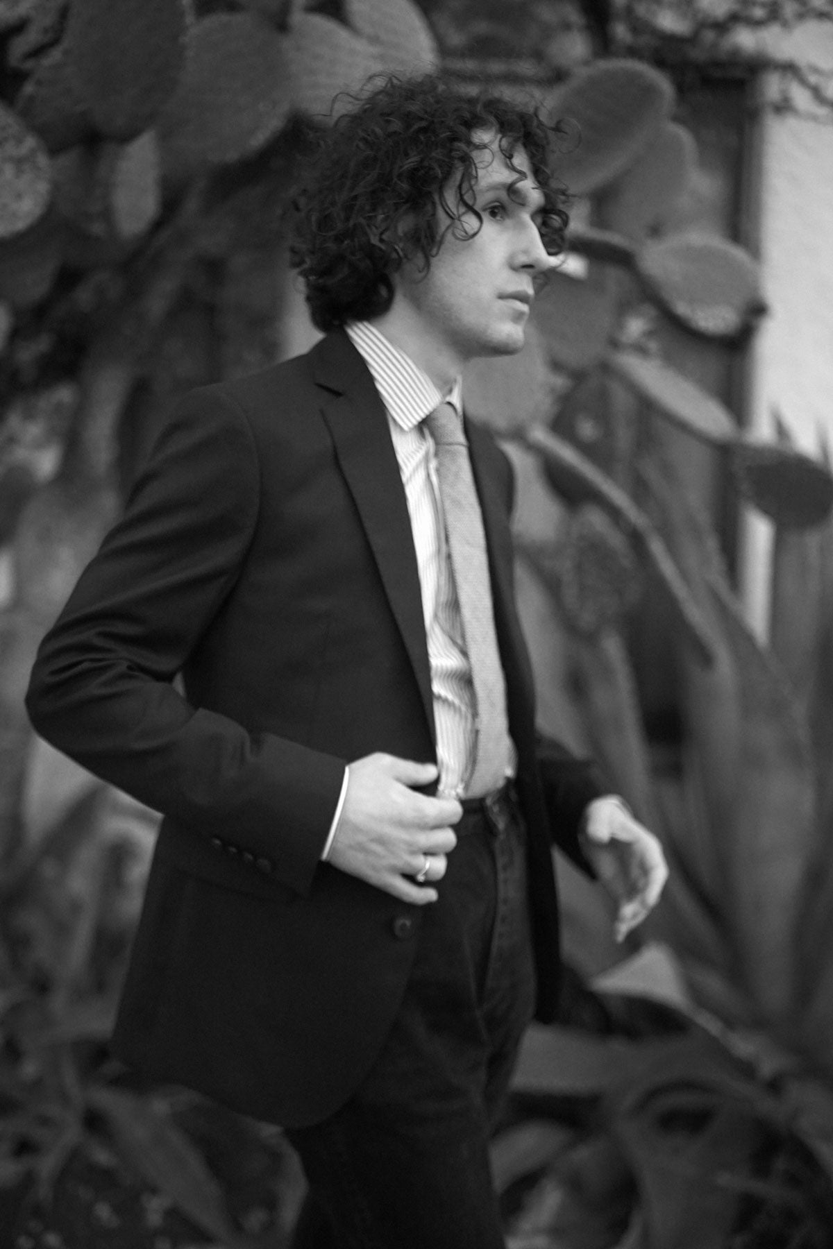 Black and white image of model wearing dark suit, striped dress shirt, and tie.