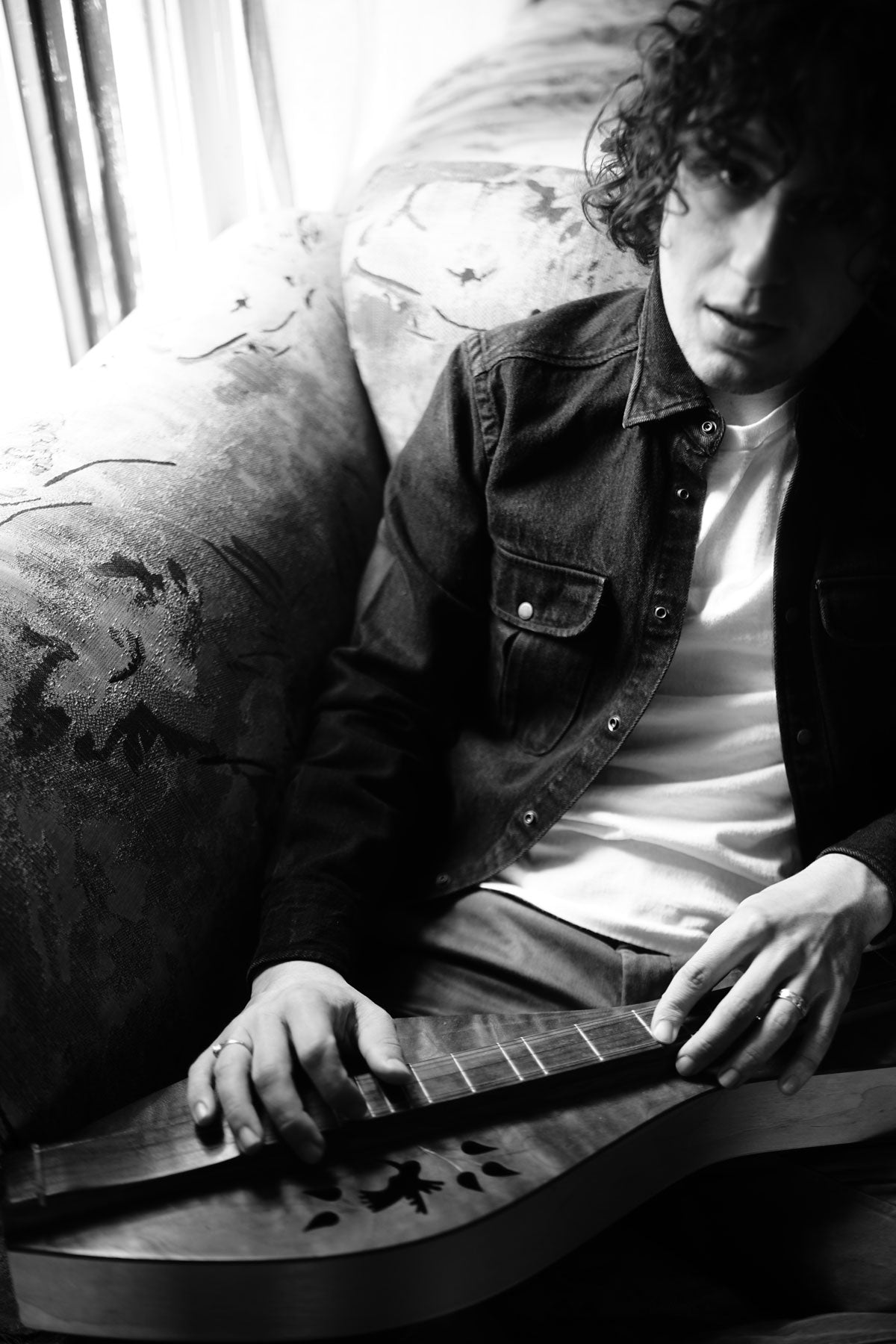 Model sits in deep armchair wearing chinos, white t-shirt, and denim overshirt. Photo is black and white.