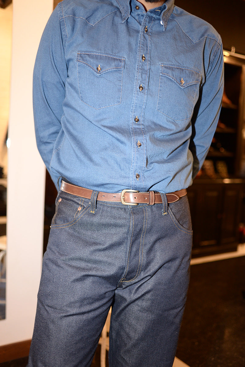 Cropped photo of a model wearing indigo denim jeans, a brown leather belt, and a lighter chambray cowboy shirt. 