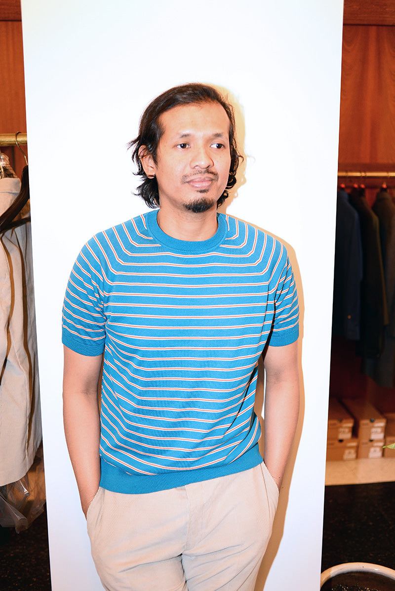 Model wears a blue and red striped t-shirt and sand colored pants. He is standing in front of a white wall. 