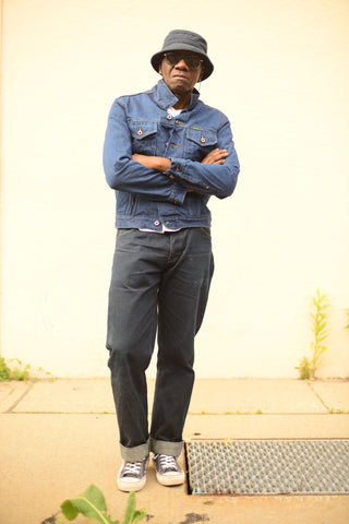 Image of Glenn wearing cuffed jeans, a denim jacket, a bucket hat, and sunglasses. His arms are crossed.