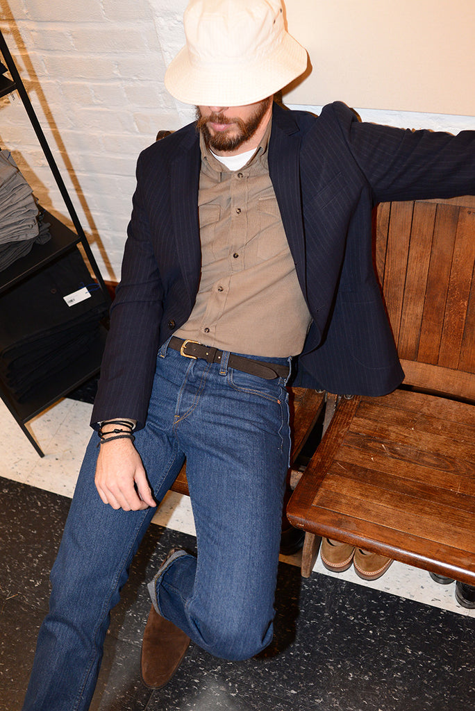 Model with a beard wears denim jeans, a tan corduroy shirt, and a navy pinstriped unstructured blazer. He is seated on a wooden bench. 