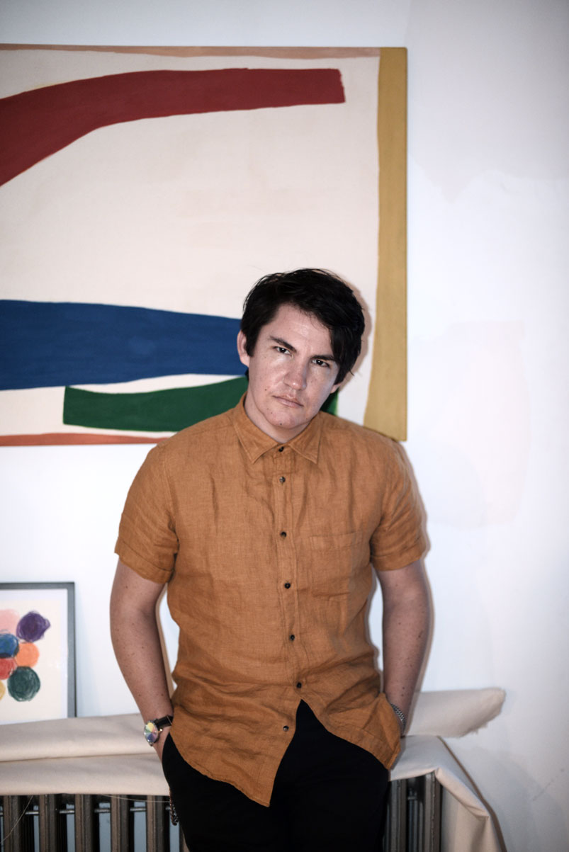 Dave wears an ochre short sleeve button down in front of one of his paintings