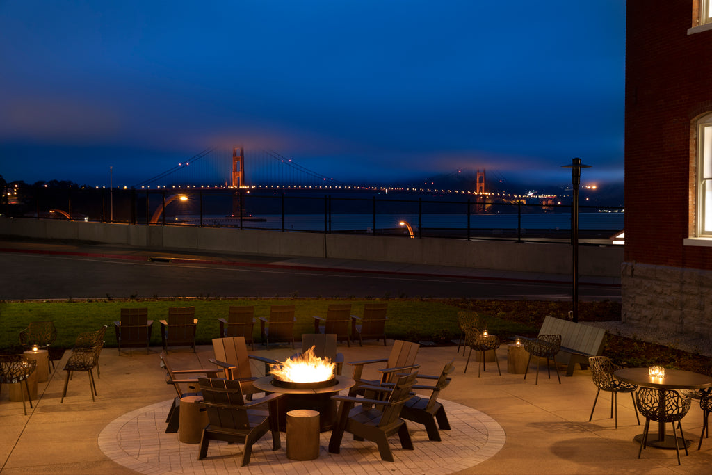 A concrete patio featuring a firepit surrounded by chairs, in the background the bay and the Golden Gate Bridge are lit by streetlights.