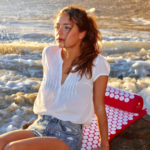 Young Woman Enjoying Bed of Nails Acupressure Mat & Pillow at the Beach with Waves behind her